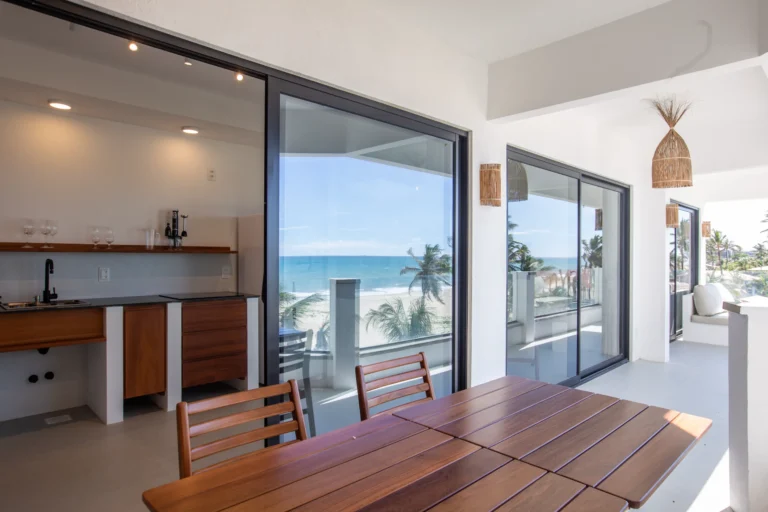 Open-plan kitchen and teak dining table in a two-bedroom beachfront apartment at Windtown Cumbuco, with glass doors onto the terrace