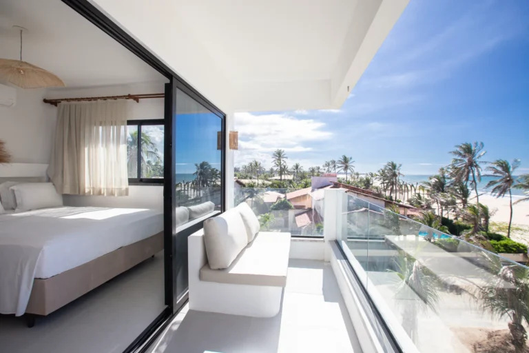 Living area of a two-bedroom beachfront apartment at Windtown Cumbuco with a white sofa and a panoramic window over the palms and the ocean