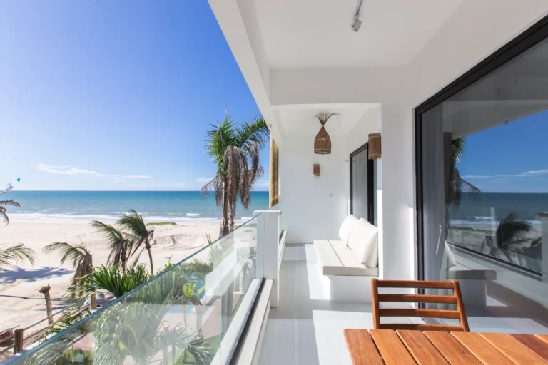 Private terrace of a one-bedroom beachfront apartment at Windtown Cumbuco, with teak seating, a glass balustrade and a view through the palms to the kite beach