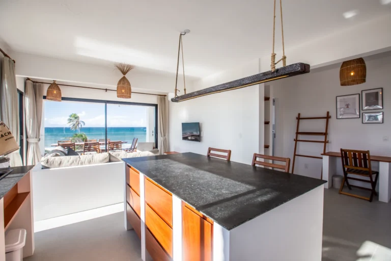Open-plan kitchen island and dining area in a three-bedroom beachfront apartment at Windtown Cumbuco, with the ocean visible through the window