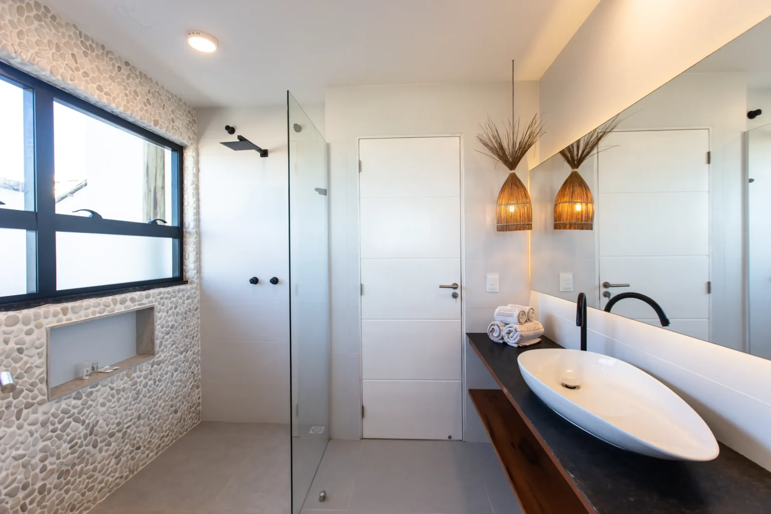 Modern bathroom in a beachfront apartment in Cumbuco with a pebble stone feature wall, glass walk-in shower, an oval vessel sink on a dark stone vanity and rattan pendant lights