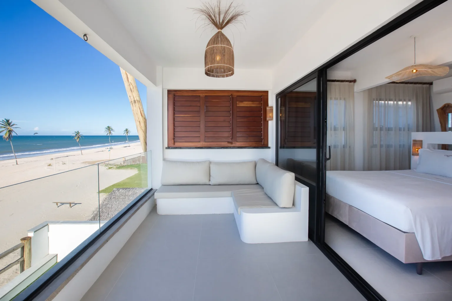 Beachfront apartment lounge area in Cumbuco with a built-in white sofa, rattan pendant light, wooden shutters and a direct ocean view through the glass balustrade