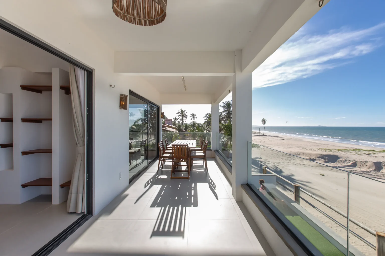 Covered terrace of a beachfront apartment in Cumbuco with a long teak dining table, built-in shelving and a glass balustrade overlooking the beach, with a kitesurf kite above the Atlantic