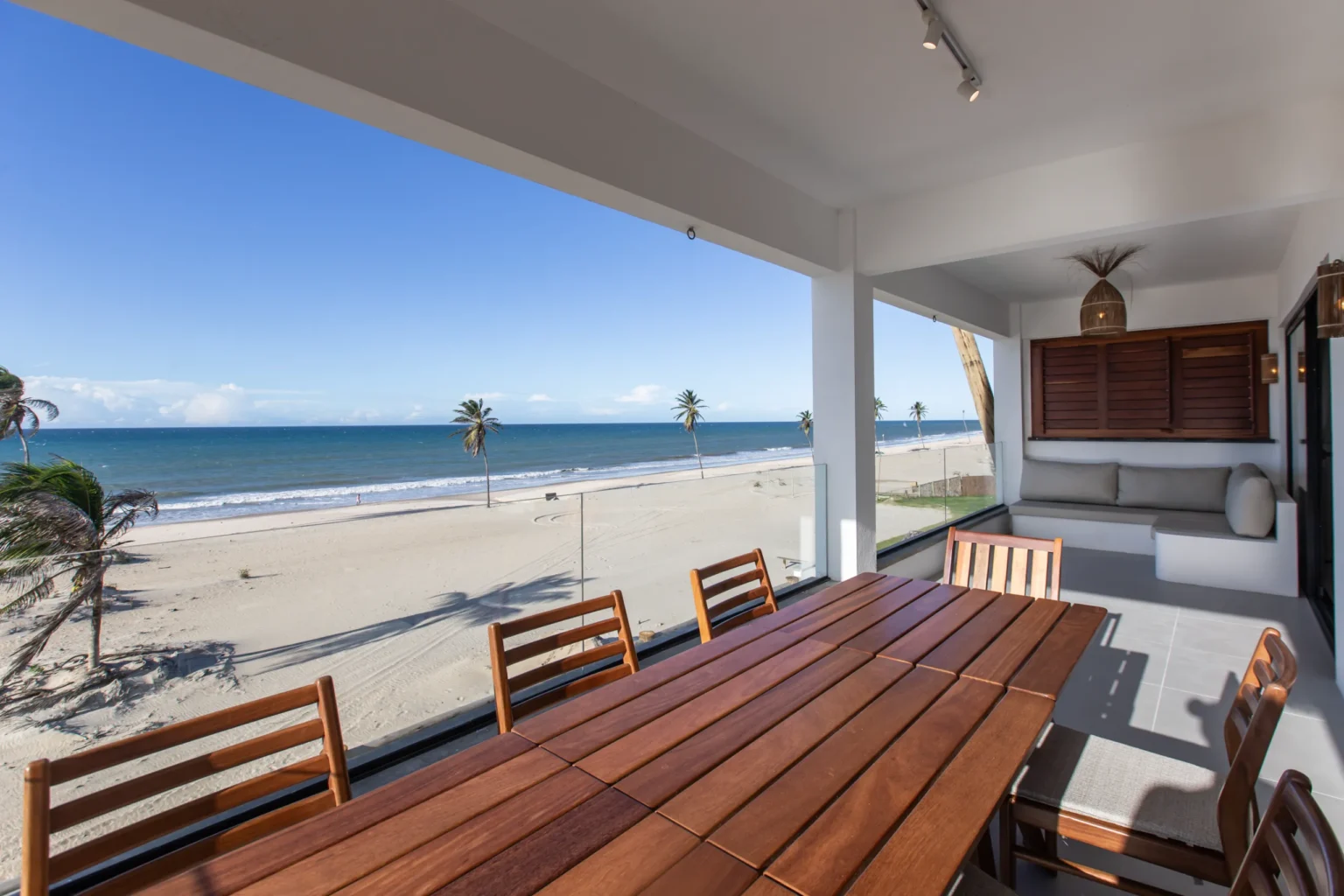 Covered outdoor dining terrace with a teak table and chairs, overlooking a white-sand beach with palm trees and calm ocean in Cumbuco, Brazil