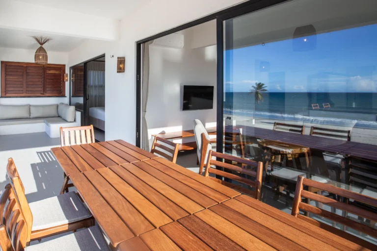 Teak dining table and bench in a three-bedroom beachfront apartment at Windtown Cumbuco, beside full-height doors onto the terrace