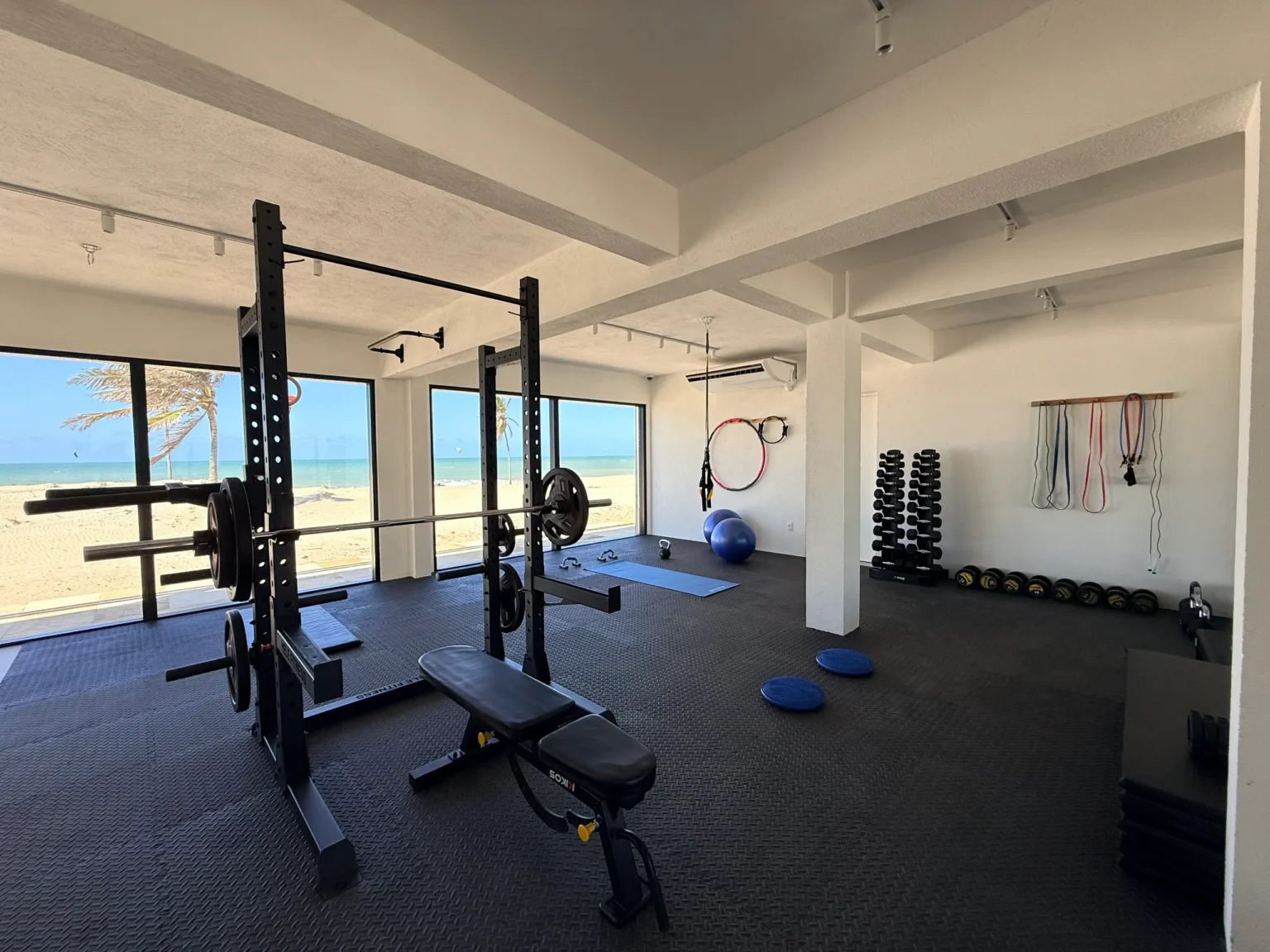 Private gym at Windtown Apartments Cumbuco with a squat rack, free weights and exercise mats in front of floor-to-ceiling windows looking onto the kite beach